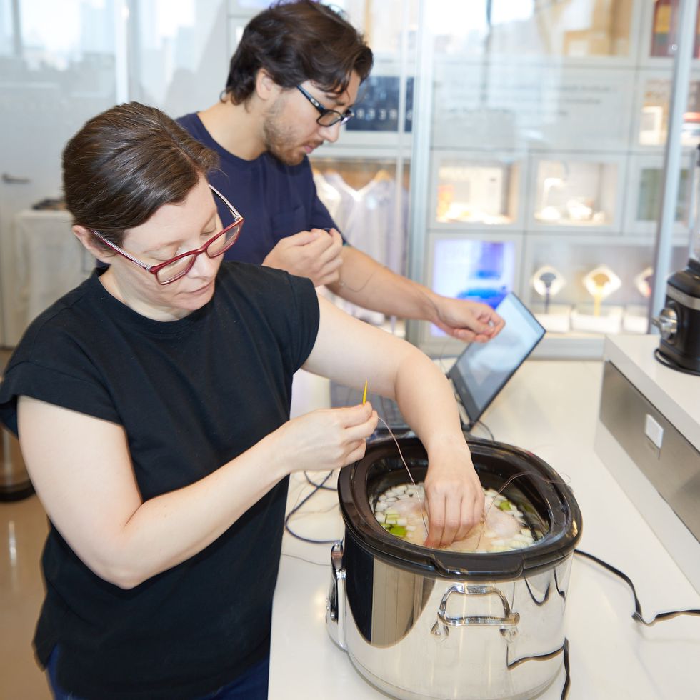 two good housekeeping institute staffers in the kitchen appliances lab one is adding thermocouples to a whole chicken in a slow cooker while the other sets up the temperature tracking software on a laptop