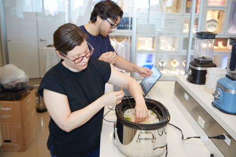 two good housekeeping institute staffers in the kitchen appliances lab one is adding thermocouples to a whole chicken in a slow cooker while the other sets up the temperature tracking software on a laptop
