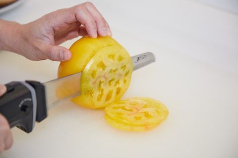 a hand holding a yellow heirloom tomato while it's being sliced by an electric knife
