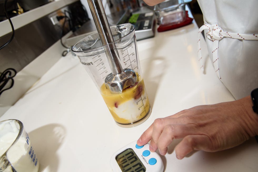 a tester poised to start a timer with one hand and in the other she holds an immersion blender in a blending cup filled with smoothie ingredients