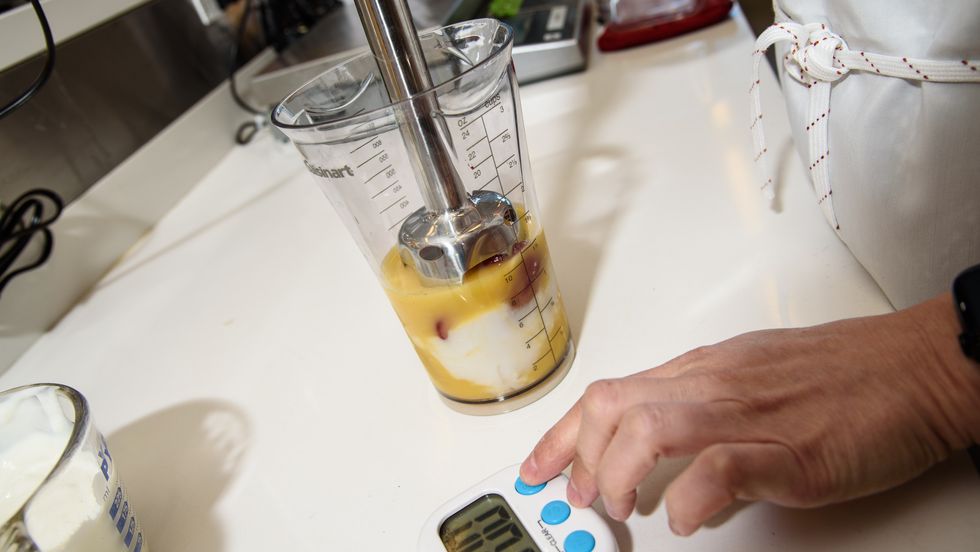 a tester poised to start a timer with one hand and in the other she holds an immersion blender in a blending cup filled with smoothie ingredients