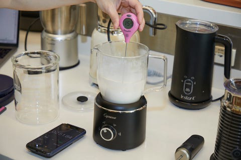 a hand holding a digital thermometer in a carafe of frothed milk on a counter with other milk frothers and a smartphone showing a stopwatch