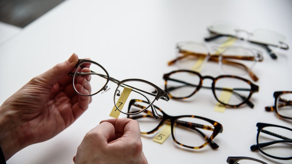 a person's hands holding a pair of glasses labeled with a number in front of a table full of labeled glasses, testing the best prescription glasses online