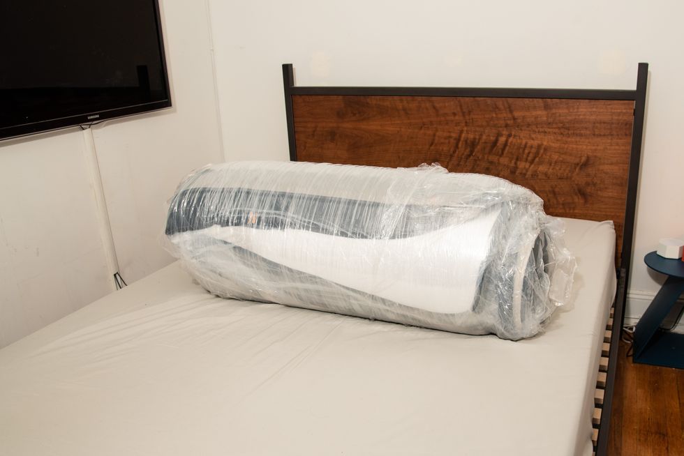 a compressed, rolled up mattress wrapped in plastic on a bed base