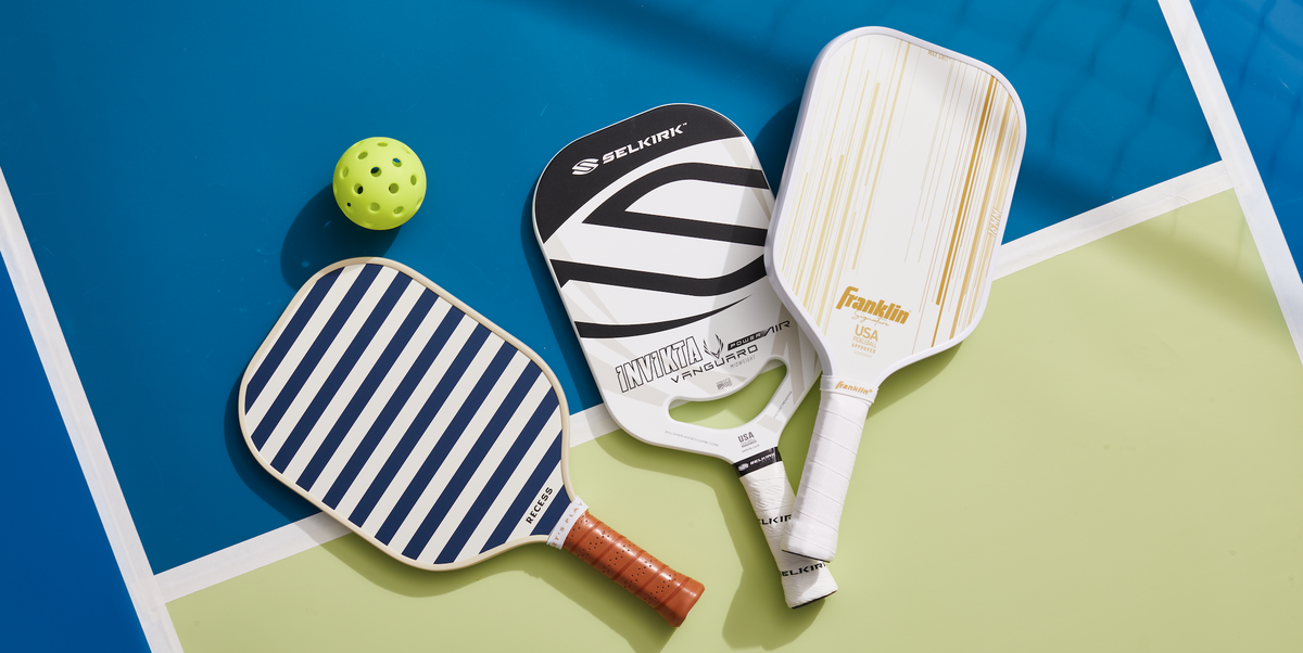 Three pickleball paddles and a ball lying on a pickleball court.
