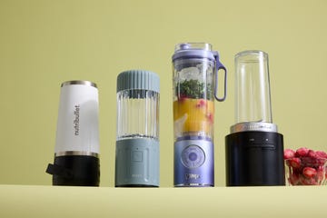 four portable blenders side by side with a bowl of frozen strawberries and smoothie ingredients in one blender