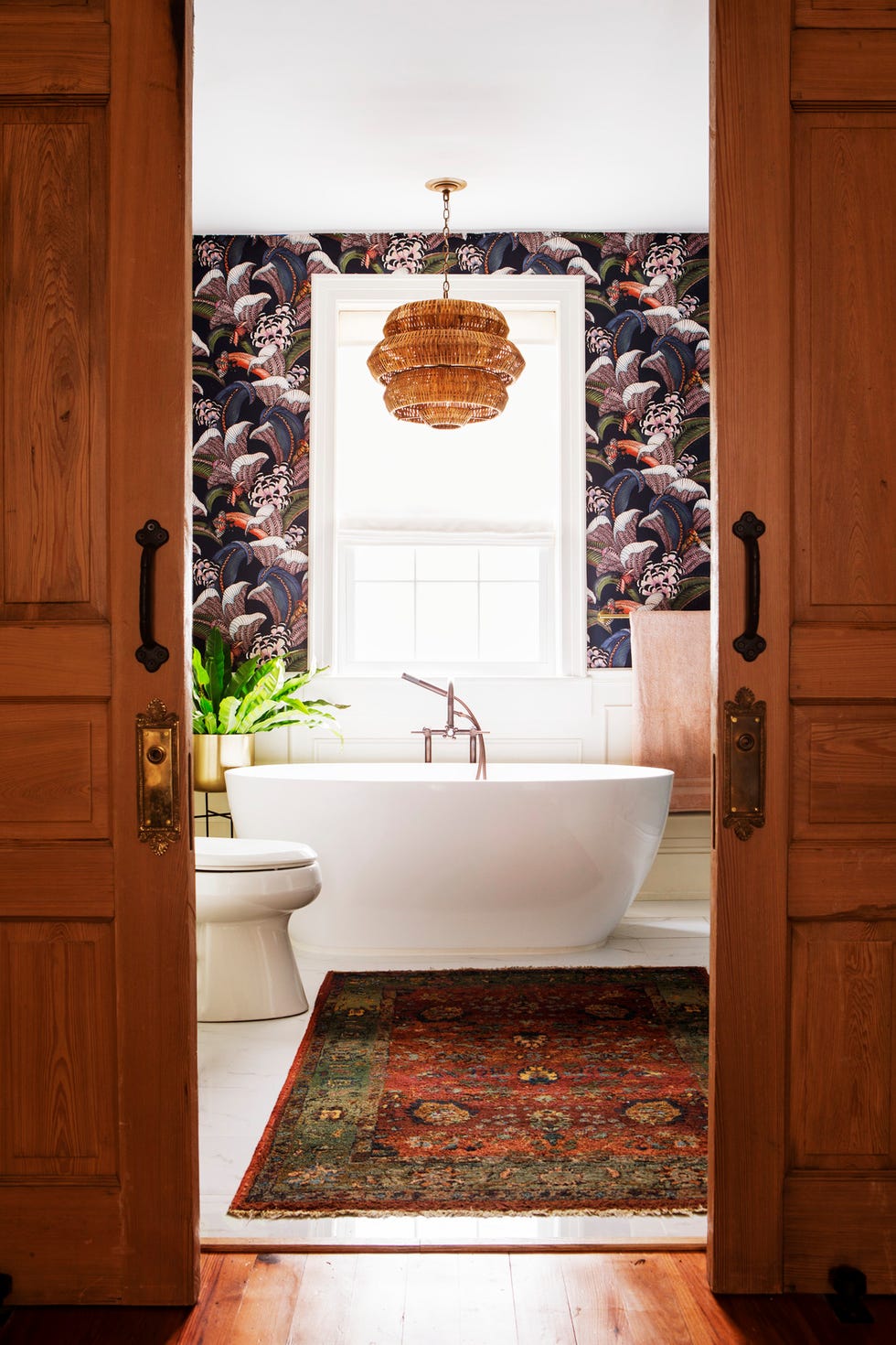 bathroom designed by hattie collins of hattie sparks interiors bold floral wallpaper, rattan lampshade, visual interest, vintage wool rug, bathtub, bright