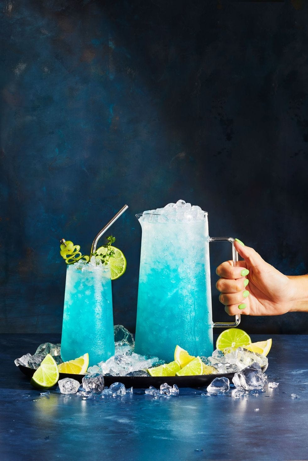 a pitcher of a blue cocktail with lime wedges as garnish