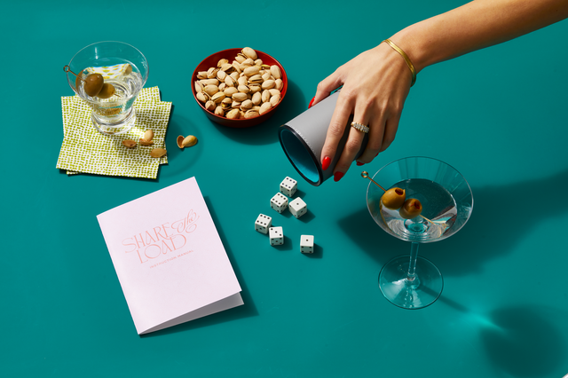 female hand shaking a dice out of a container surrounded by cocktails and snacks