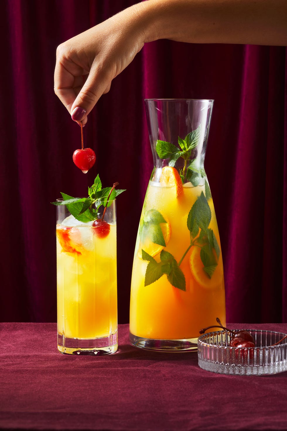 orange colored punch in a carafe garnished with mandarin slices and mint alongside a tall glass of the punch