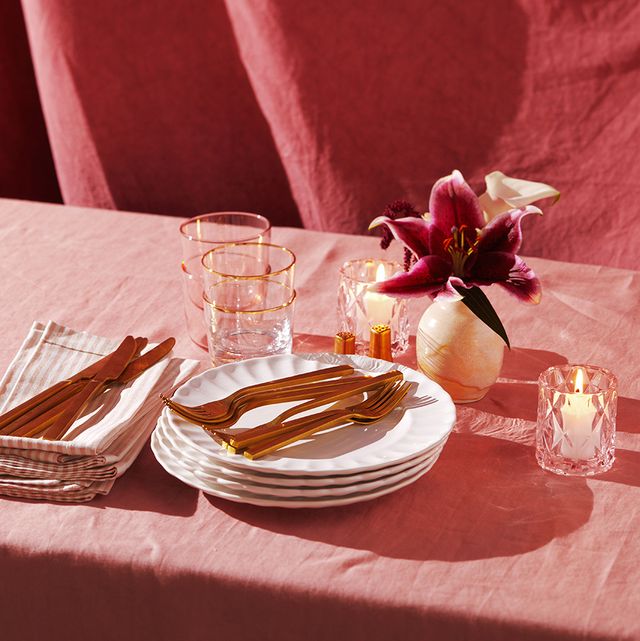 stack of white scalloped plates and gold utensils on pink table cloth with lily in vase and candles