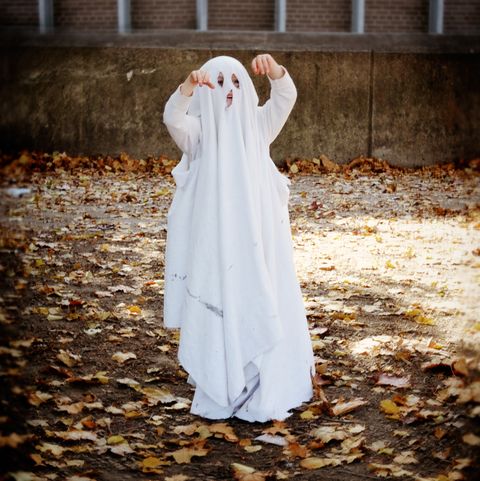 color portrait of girl wearing sheet with eye and mouth holes as halloween ghost costume, standing outside surrounded by fallen leaves