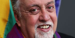 gilbert baker smiles at the camera, behind him is a rainbow flag and he wears a rainbow scarf with his black top