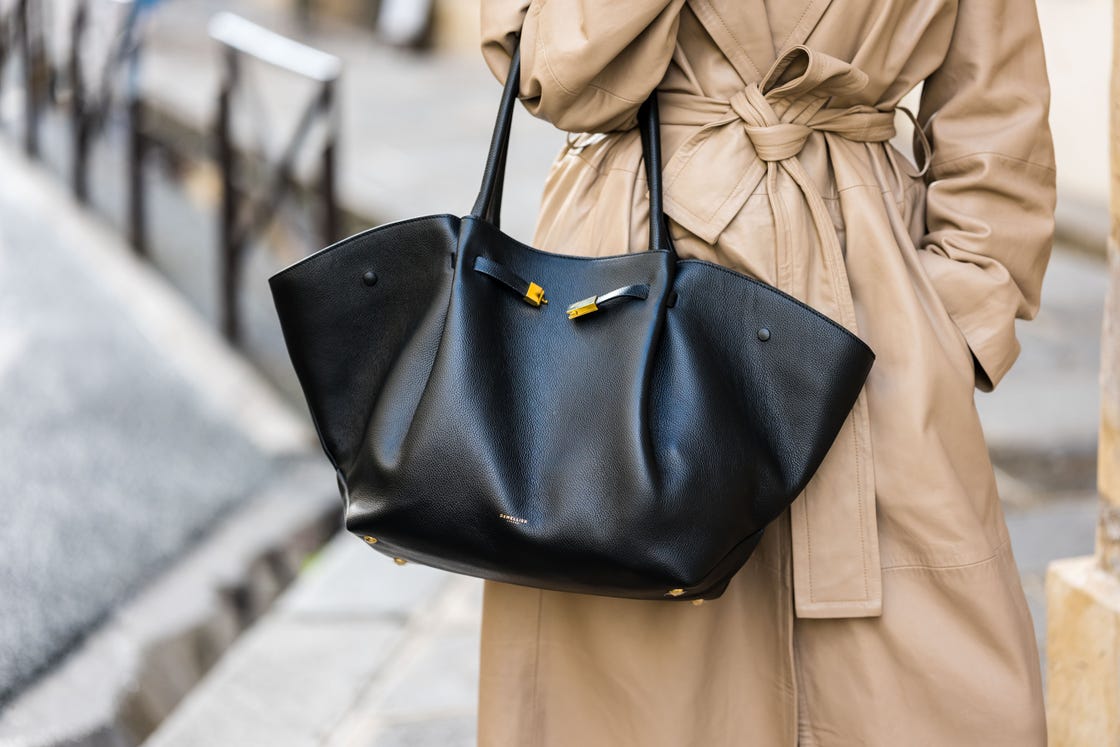 a woman carry a black leather tote bag in teh crook of her elbow with a tan trench coat on the street in paris in a roundup of the best designer travel bags 2024