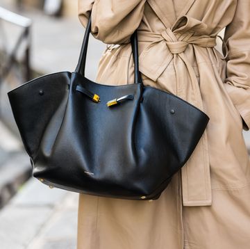 a woman carry a black leather tote bag in teh crook of her elbow with a tan trench coat on the street in paris in a roundup of the best designer travel bags 2024