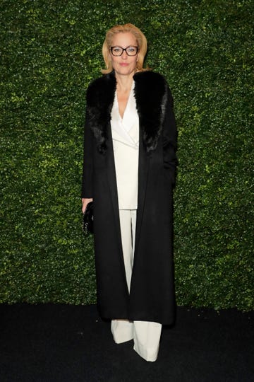 gillian anderson, wearing a black winter coat, white suit and tortoiseshell glasses, stands in front of a 'living wall' at a party