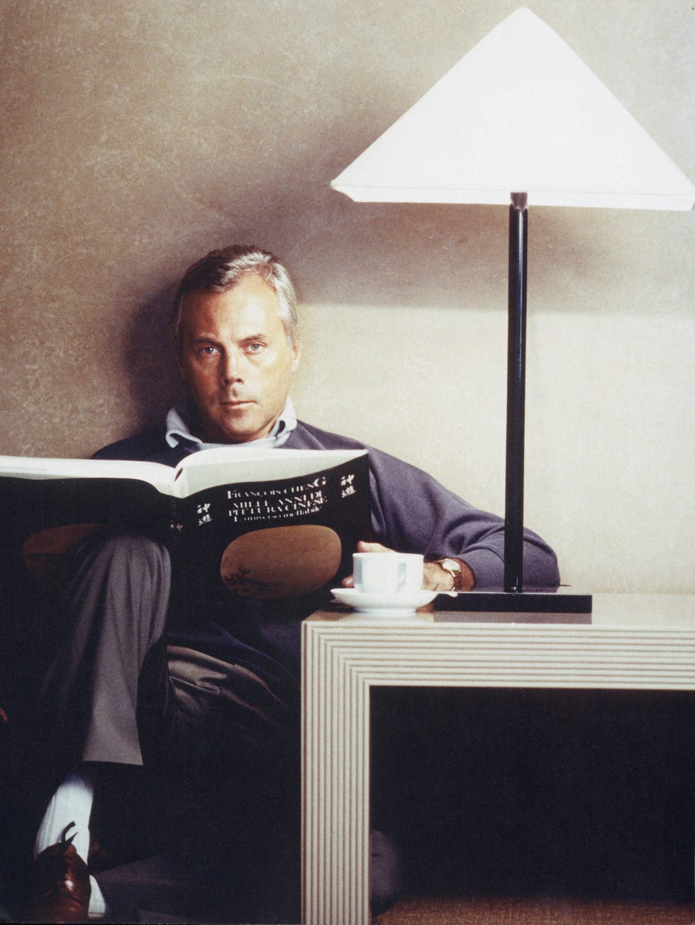 a person reading a book while seated beside a lamp and a cup