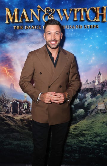 giovanni pernice, a man stands clasping his hands and smiling at the camera, he has short dark brown hair and light facial hair, he wears a black shirt and trousers with brown blazer jacket