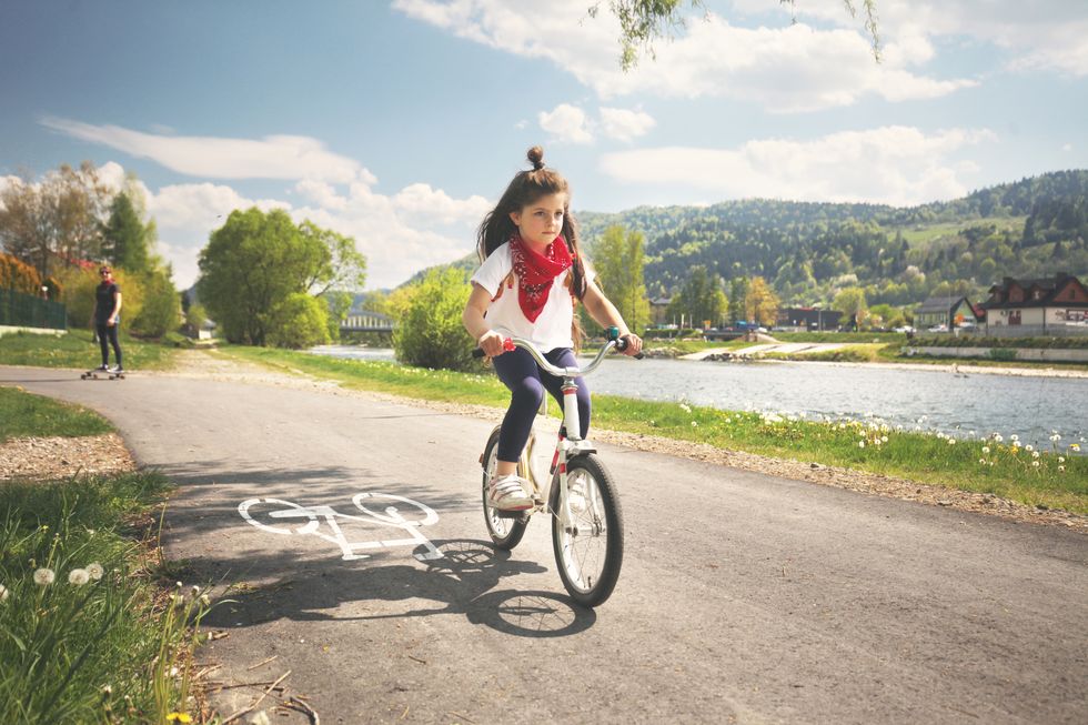 girl riding bicycle on road along river, mother on longboard in background