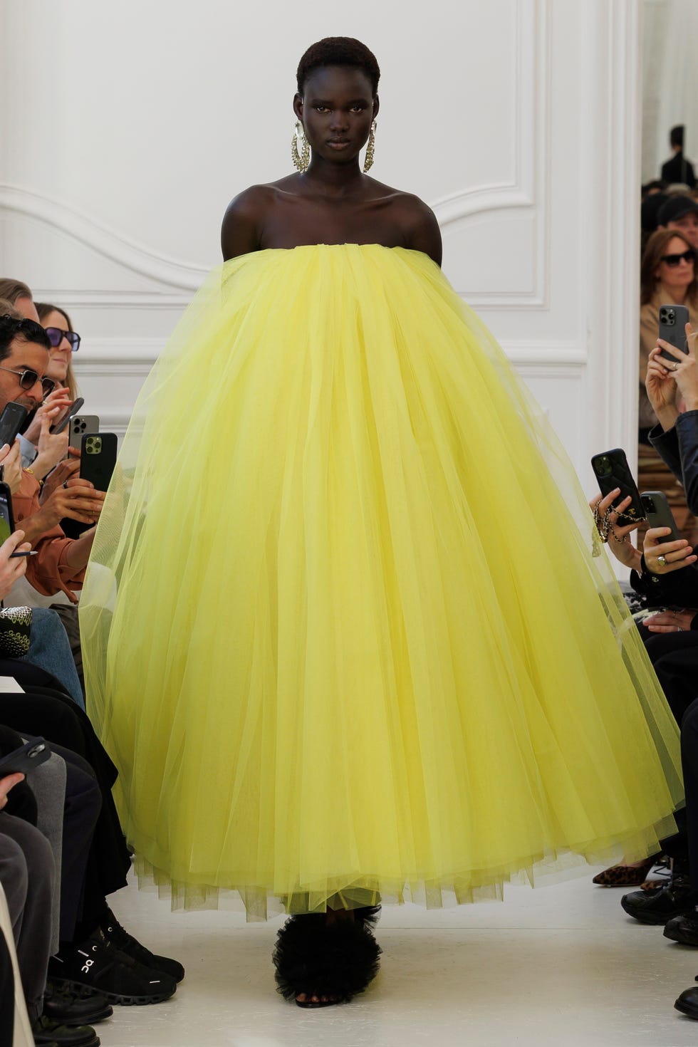 Fashion model wearing a vibrant yellow gown surrounded by audience members with phones