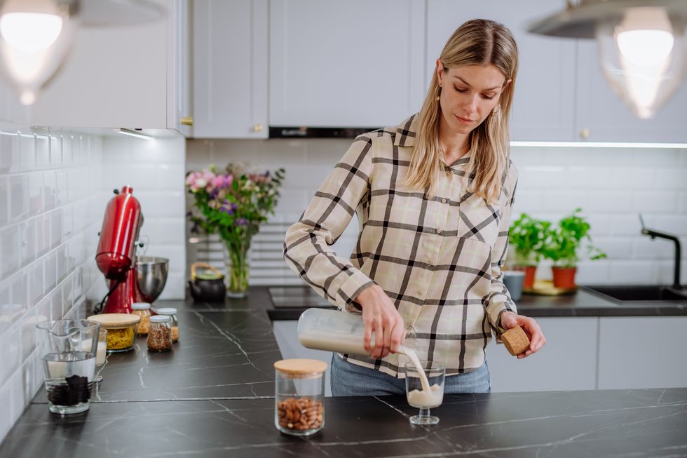 Μια γυναίκα ρίχνει γάλα αμυγδάλου σε ένα ποτήρι στην κουζίνα, έννοια υγιεινού vegan προϊόντος