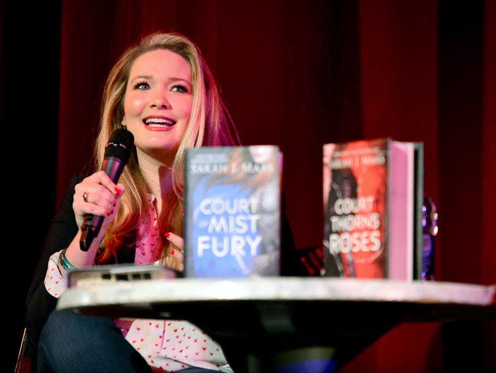 sarah j maas holds a microphone and speaks while sitting behind a table in the foreground, on the table are two of her books, one with a blue cover, one with a red cover, she wears a patterned white and red blouse with jeans