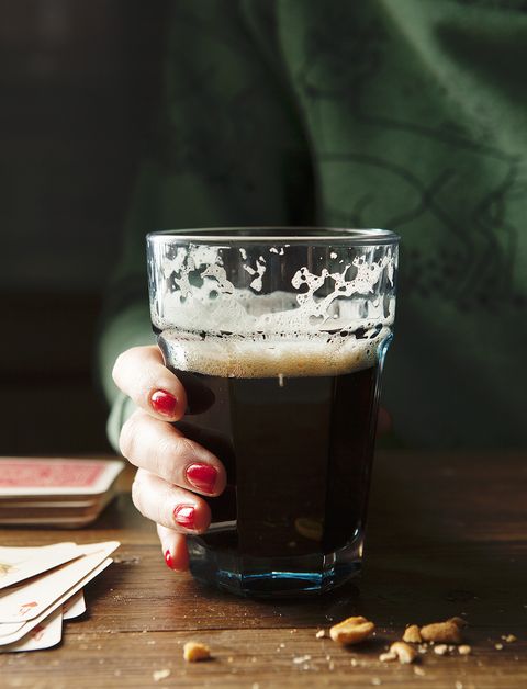 glass of drunk guinness with a ladies hand with red nails and a green sweater holding glass at a wooden table surrounded by nuts and playing cards