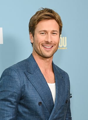 glen powell smiles at the camera while standing in front of a light blue background, he wears a blue suit jacket and white shirt