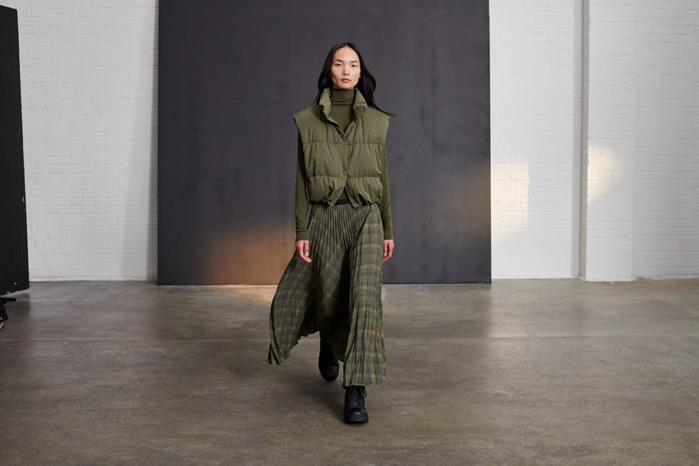 a person wearing a green vest and a long plaid skirt walking in a minimalist studio setting