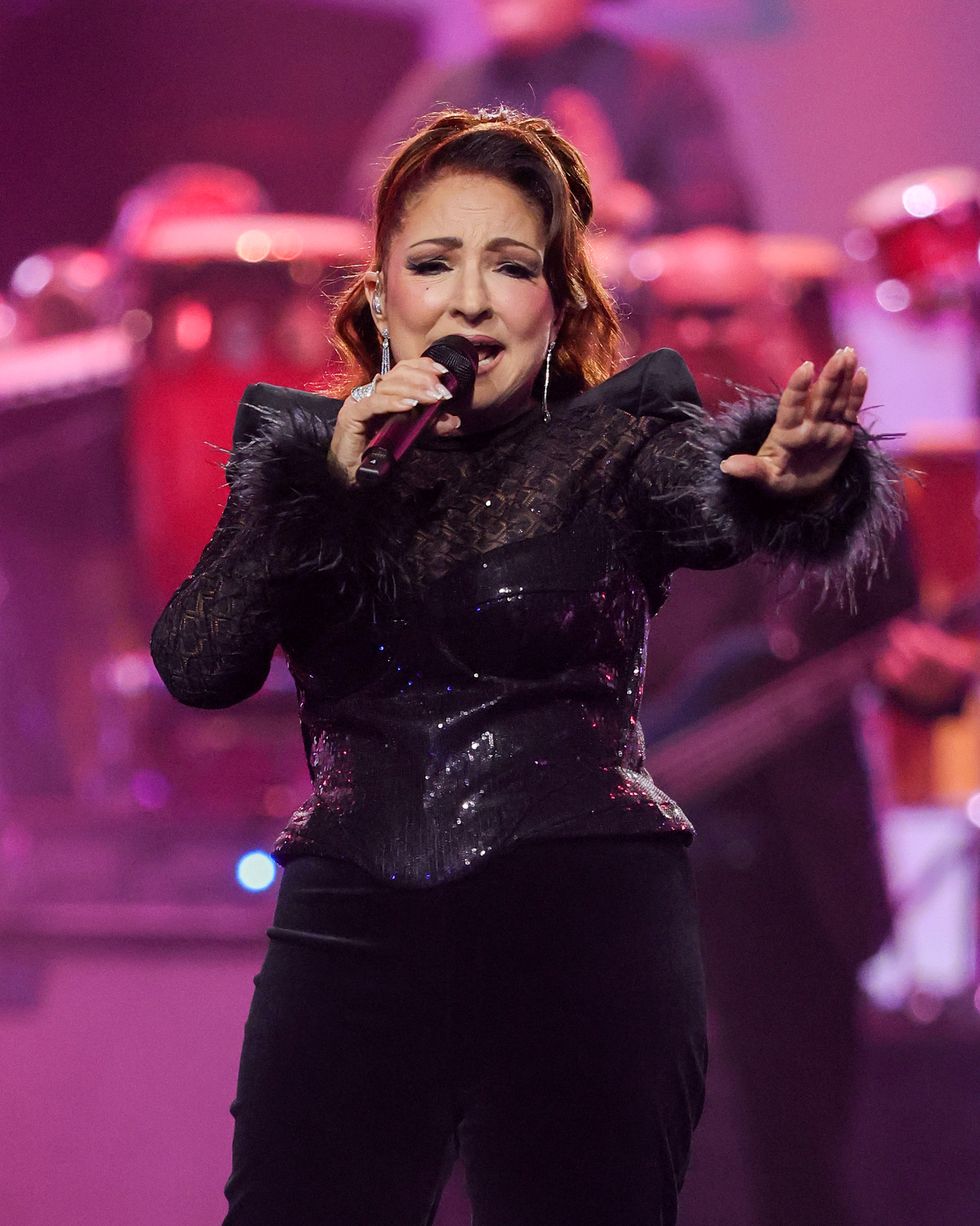 a woman sings into a wireless microphone she holds with one hand while holding out her other hand, she wears all black and stands on a pink lit stage