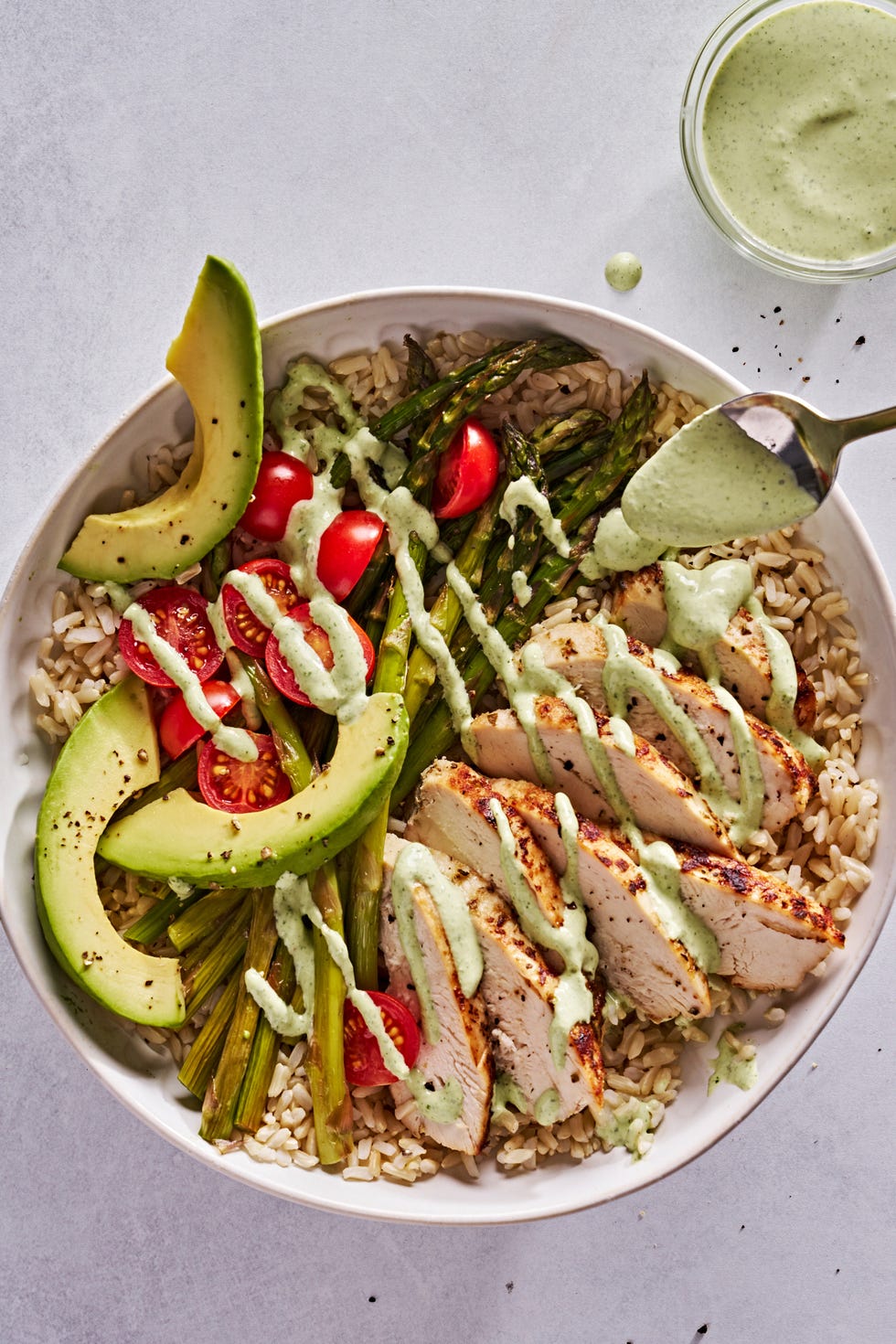 bowl of brown rice topped with chicken, sliced baby tomatoes, avocado slices, roasted asparagus, and green goddess dressing