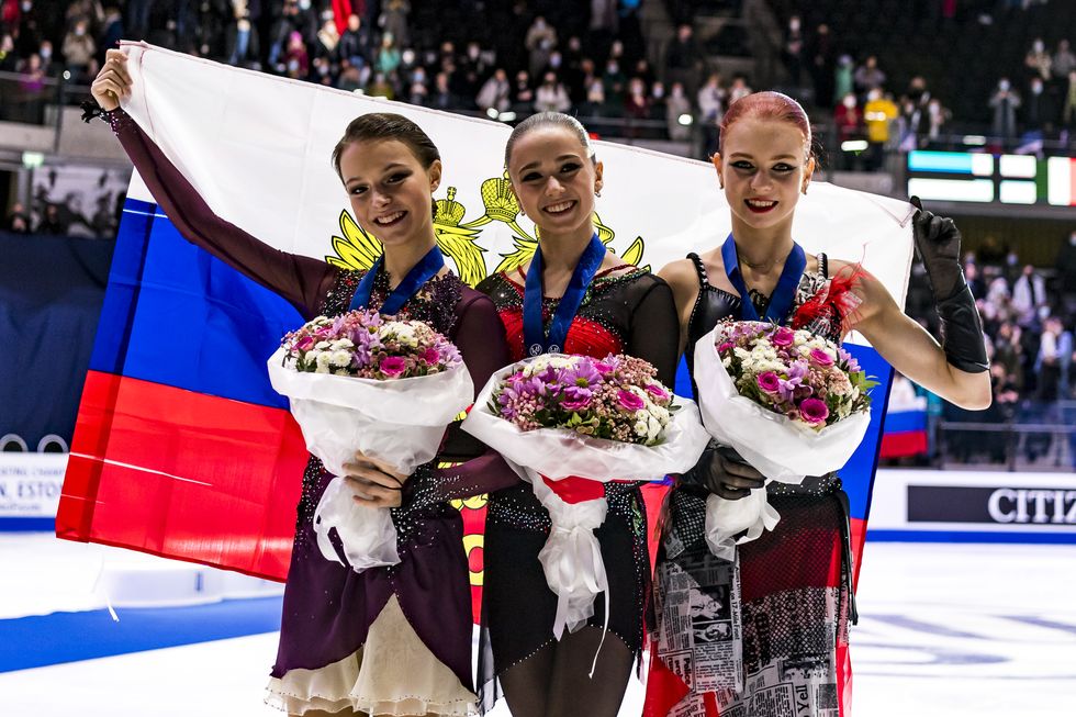 kamila valieva, alexandra trusova, and anna shcherbakova all smiling after sweeping the podium at a competition while holding the russian flag behind them