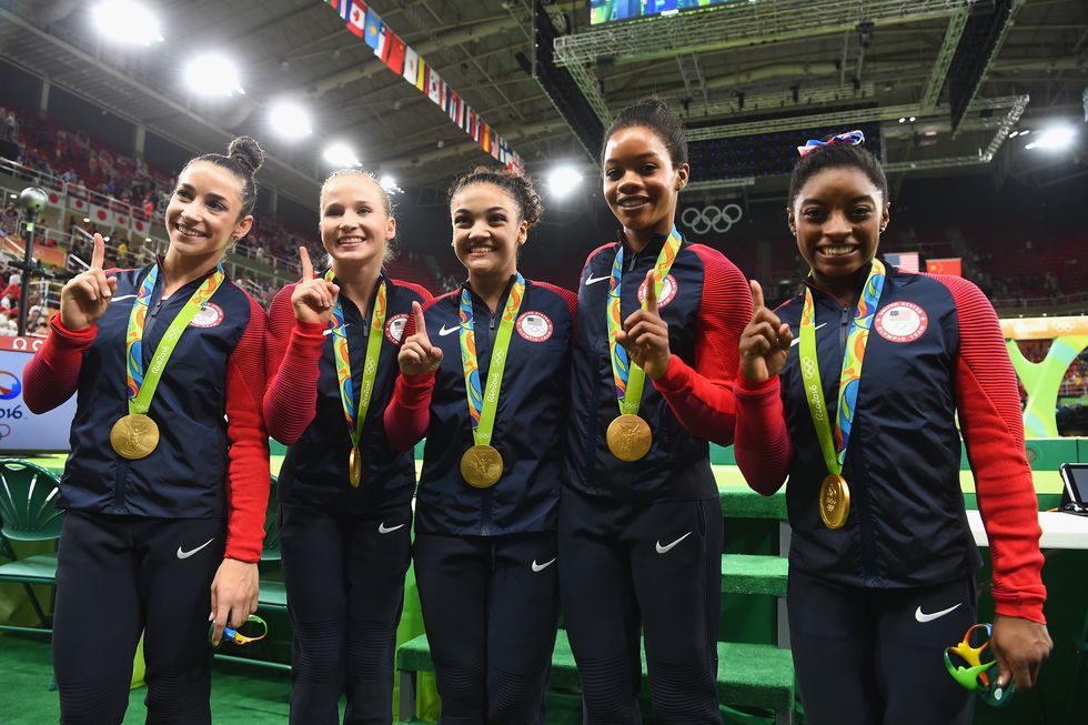 ali raisman, madison kocian, lauren hernandez, gabby douglas, and simone biles stand together and smile for photos, each wears a gold olympic medal and points one finger into the air, they have on navy blue and red team usa sweats