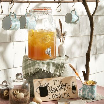 golden gobbler cocktail in a big pitcher jar on a table with a sign and cups hanging above from a wooden dowel