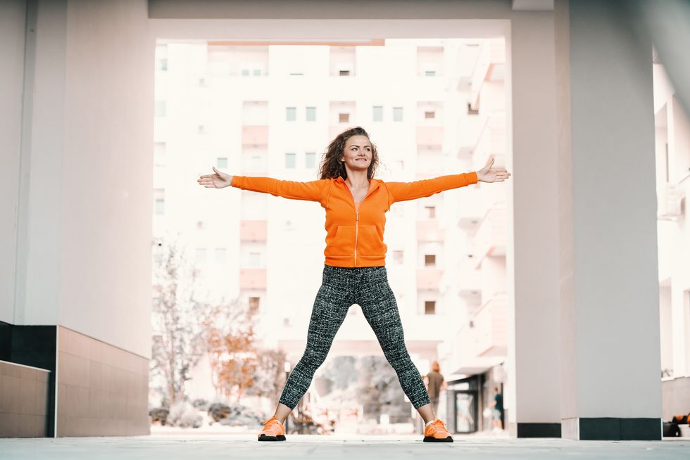 good looking fit healthy smiling caucasian woman in sportswear and with curly hair doing jumping jacks in passage on sunny day
