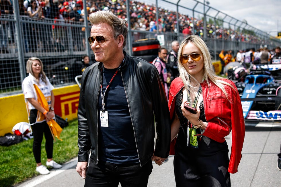 gordon ramsay and tana hutcheson walk hand in hand on a race car track, they wear jackets and sunglasses, race cars and stands are behind them