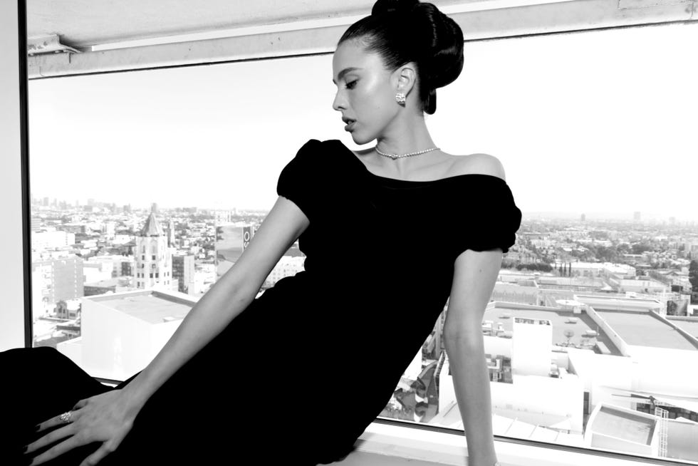 elegant woman in a black offshoulder dress posing by a window with a city skyline view