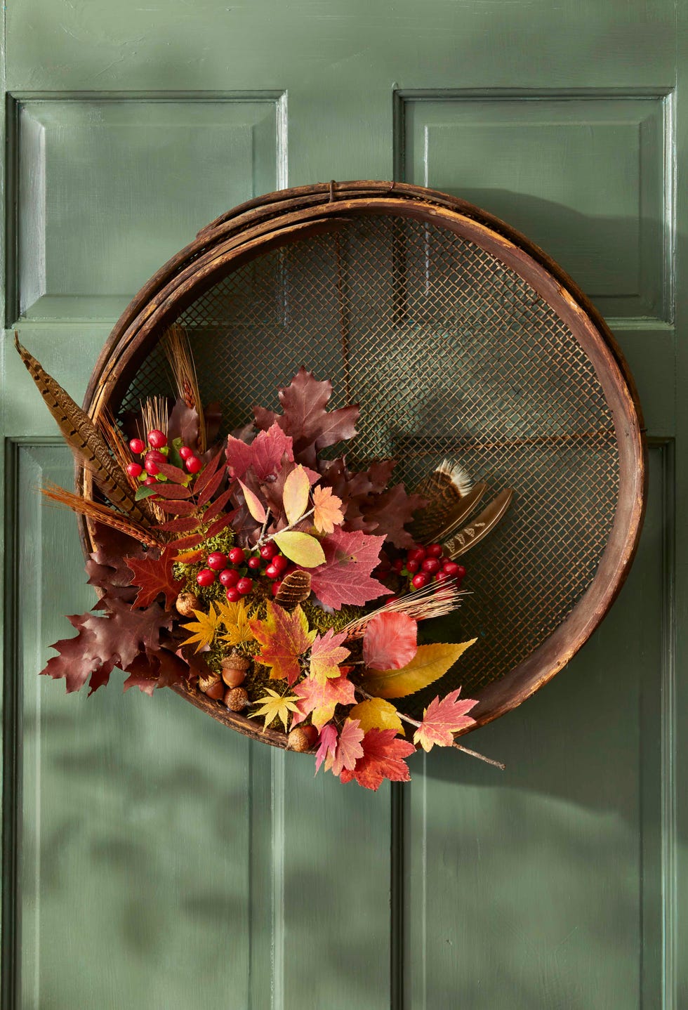 a wreath made from a vintage grain sifter with leaves, feathers, and acorns hung on a green door