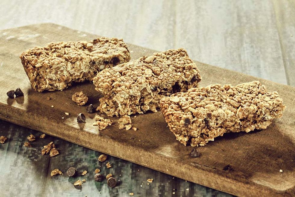 three home made choc chip granola and oats bars on a wooden board sitting on a back lit wood texture surface with additional choc chips and oats crumbs next to it