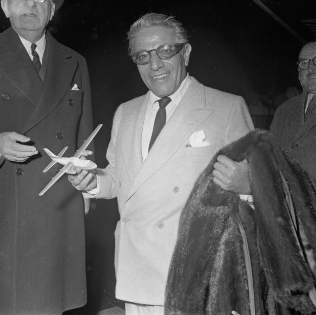 aristotle onassis smiles at the camera, he wears a suit jacket, collared shirt, tie, and glasses