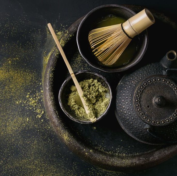 green tea matcha powder and hot drink in black bowls standing with iron teapot, bamboo traditional tools spoon and whisk in terracotta tray over dark metal background top view with space