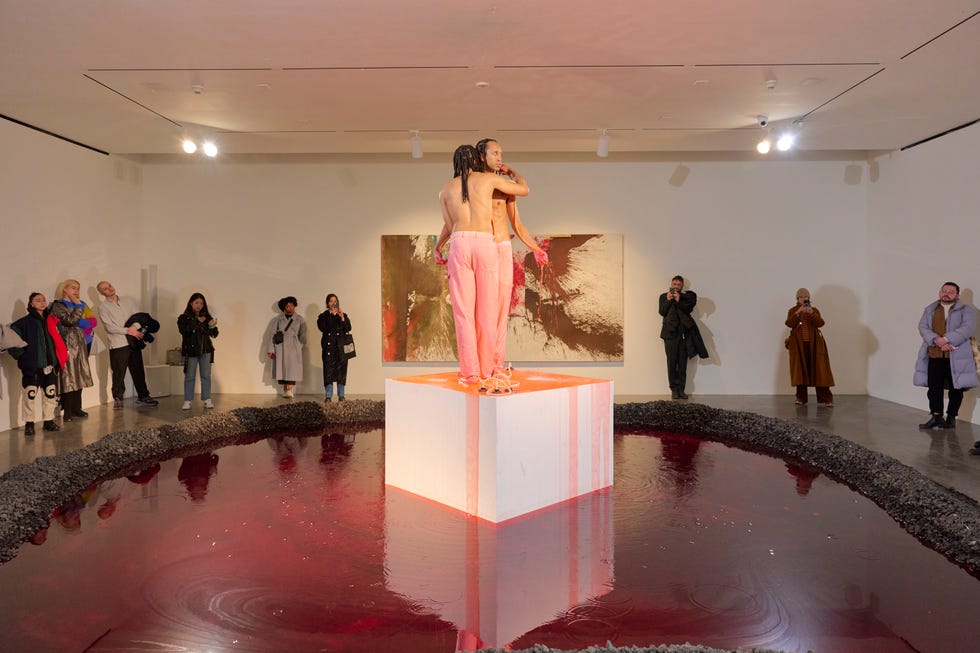 exhibition featuring a performance on a pedestal surrounded by red liquid and an audience