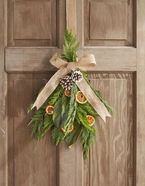 swag wreath made from greenery, dried fruit, and pine cones hung on a wood door and embellished with a burlap ribbon