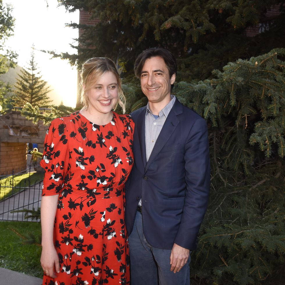 greta gerwig and noah baumbach pose for a photo together while standing in front of an evergreen tree, both smile and gerwig looks right of the camera, she wears a red dress with a black and white floral pattern, he wears a navy suit jacket, gray collared shirt and navy pants