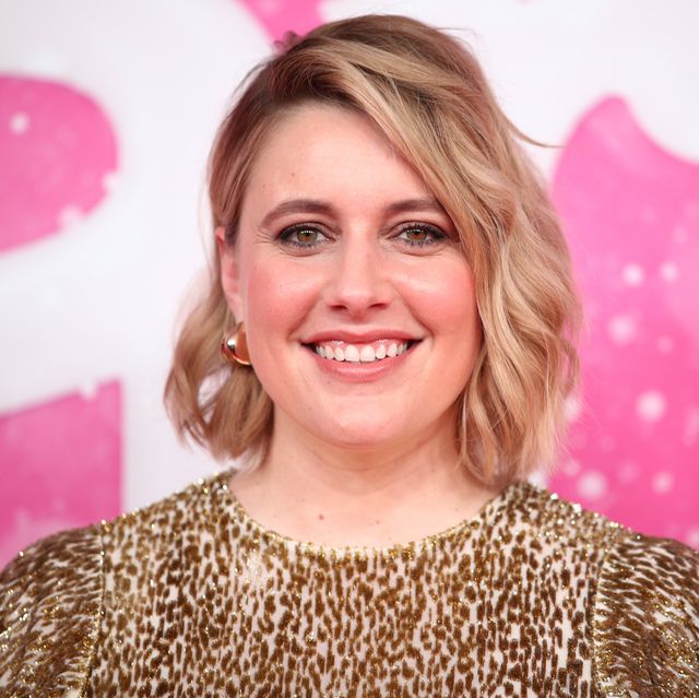 greta gerwig smiles at the camera, she wears a gold and white dress with gold hoop earrings, her shoulder length hair is styled down and she has on makeup