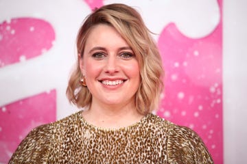greta gerwig smiles at the camera, she wears a gold and white dress with gold hoop earrings, her shoulder length hair is styled down and she has on makeup