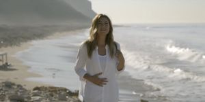 ellen pompeo, a white brunette actress, stands on a beach shooting a scene for grey's anatomy she is wearing a white button down shirt and blue jeans