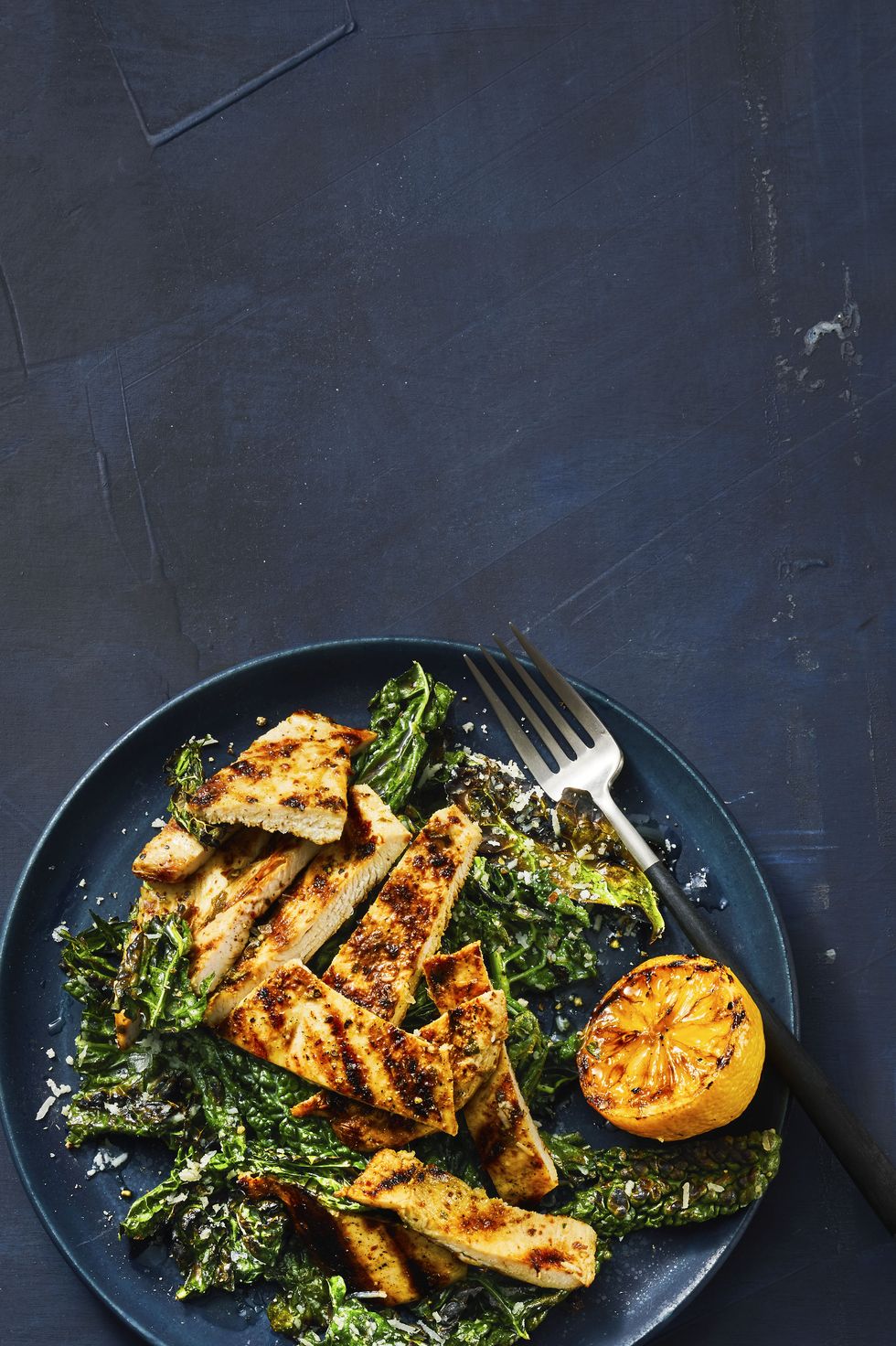 grilled lemony chicken and kale on a dark blue plate with a grilled lemon served on the side