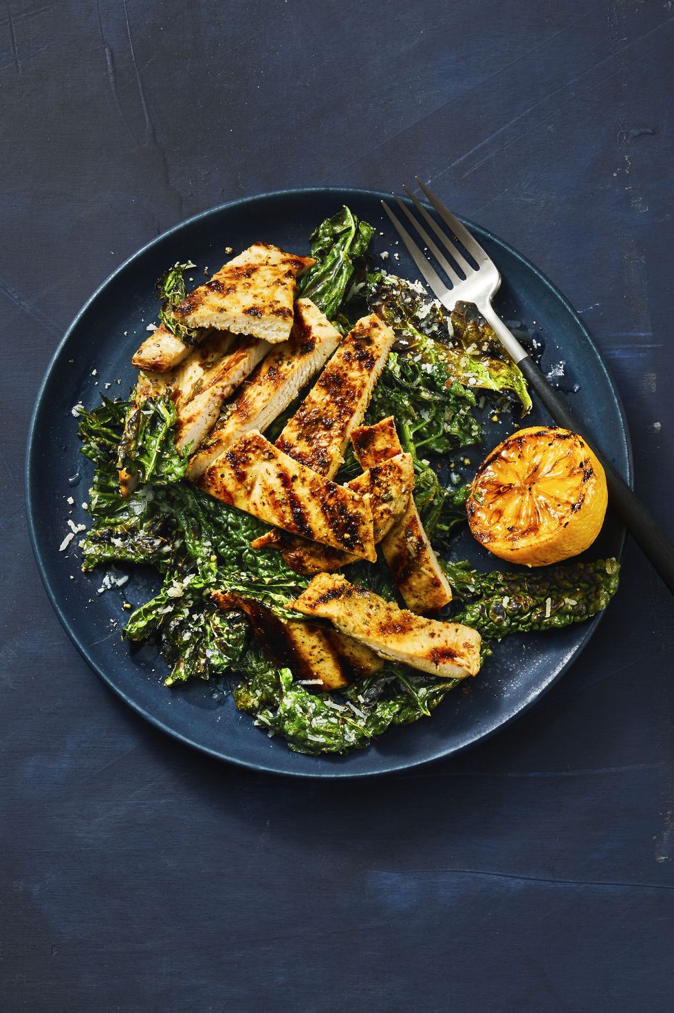 grilled lemony chicken and kale on a dark blue plate with a grilled lemon served on the side