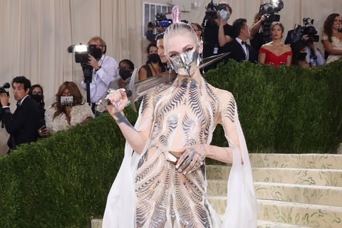 grimes, wearing a dress with black patterns and a silver facemask, holding a sword on her shoulder, standing on a stairway, with hedges separating her from several people and photographers